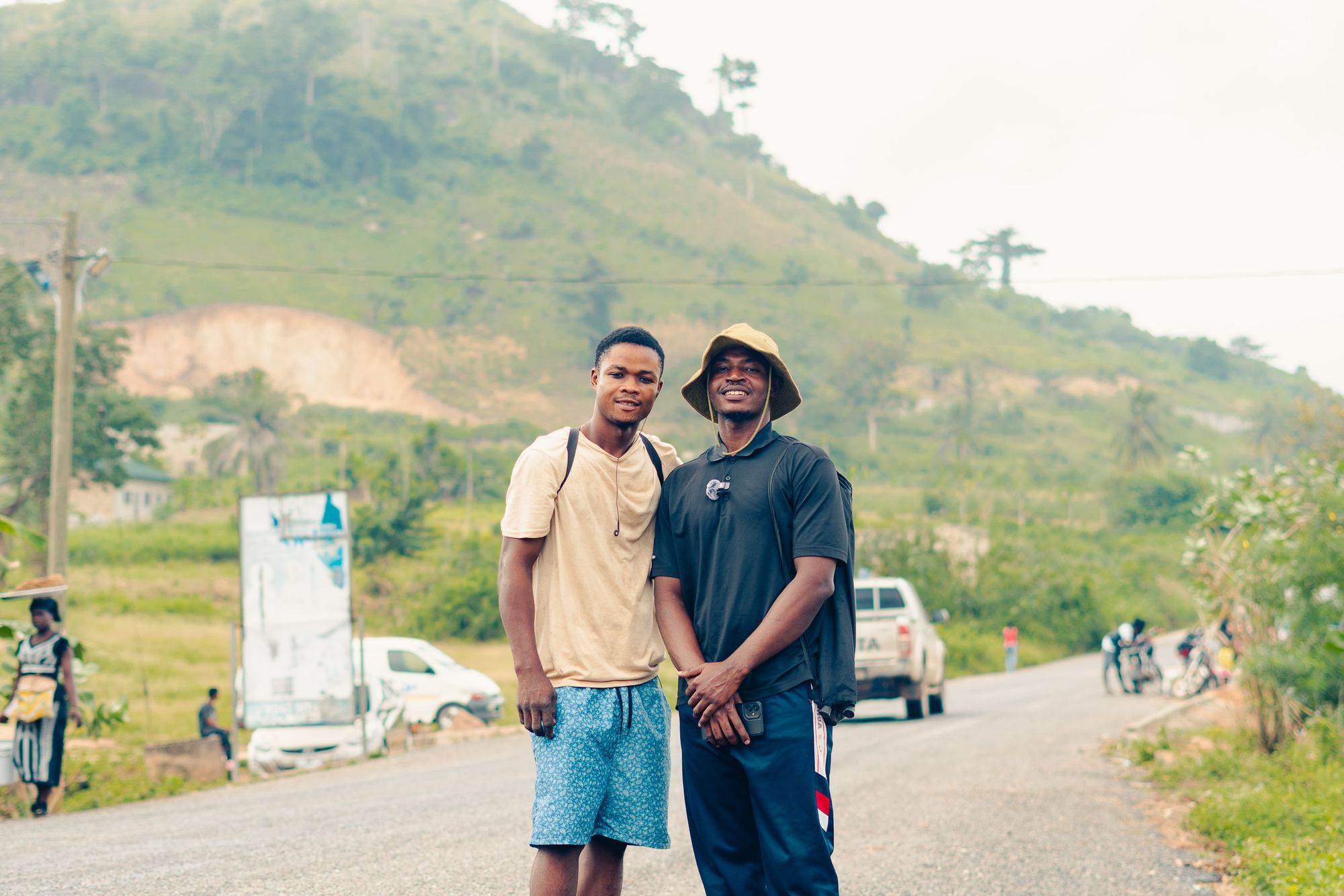 Deux amis sourient au bord d'une route, devant des collines vertes.